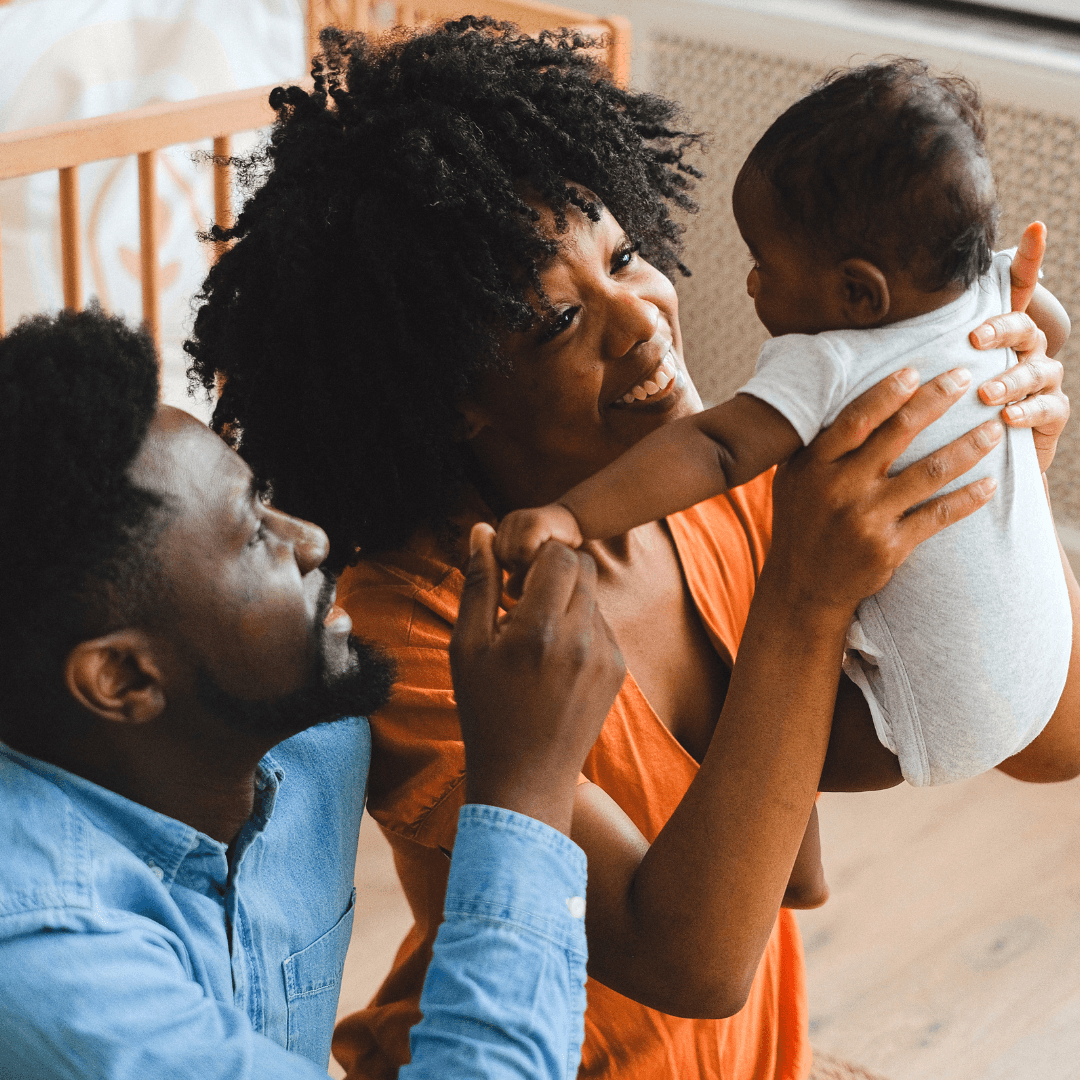 A mother lifting her baby while the father holds the baby's hand beside a wooden cot